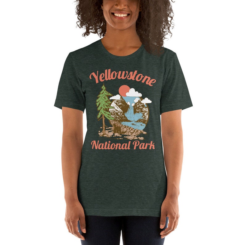 May include: A dark green t-shirt with a vintage-style illustration of a mountain scene with a waterfall, a pine tree, and a red sun. The text "Yellowstone National Park" is printed in a light pink font.