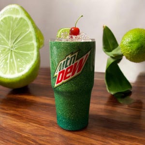 Puede incluir: Vaso verde con purpurina con un logotipo de Mountain Dew. El vaso está lleno de hielo y tiene una cereza en la parte superior.
