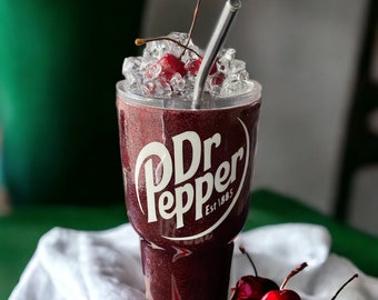 May include: A tall glass filled with a dark red beverage with ice and a cherry on top. The glass has a white Dr Pepper logo with the text "Est. 1885". A metal straw is in the glass. There are several red cherries on a white and red striped towel.