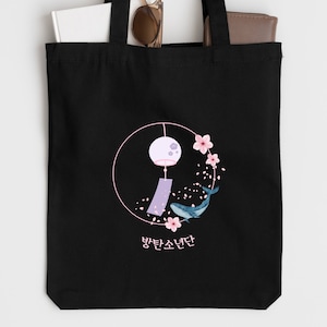 Könnte beinhalten: Schwarze Tragetasche mit einem weißen Kreisdesign, das eine rosafarbene Windspiel, rosafarbene Kirschblüten und einen blauen Wal zeigt. Der Text "방탄소년단" ist unter dem Design gedruckt.