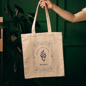 Puede incluir: Una bolsa de tela beige con un diseño gráfico en blanco y negro que presenta una serpiente y el texto "She got a hold of me like a Python".
