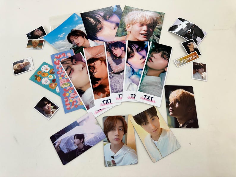 May include: A collection of photo cards featuring a K-pop boy band. The cards are printed with various images of the band members, including close-ups of their faces and full-body shots. The cards are decorated with the band's name, "TXT", and the word "Tomorrow".
