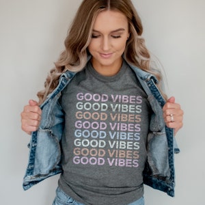 May include: A gray t-shirt with the phrase "GOOD VIBES" repeated in rainbow colors. The shirt is being worn by a person with long blonde hair and a denim jacket.
