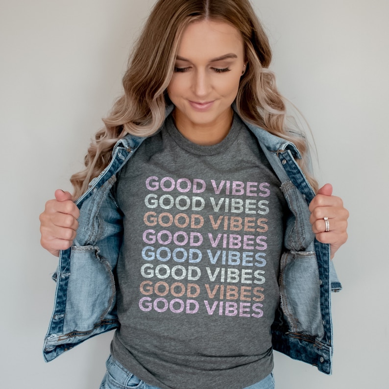 May include: A gray t-shirt with the phrase "GOOD VIBES" repeated in rainbow colors. The shirt is being worn by a person with long blonde hair and a denim jacket.
