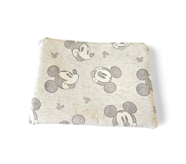 Puede incluir: Una bolsa rectangular gris claro con un patr&oacute;n repetido de caras sonrientes de Mickey Mouse y siluetas de la cabeza de Mickey Mouse en gris. La bolsa est&aacute; hecha de una tela suave y tiene un cierre de cremallera.