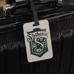 May include: A white luggage tag with the word "Slytherin" and a green serpent crest. The tag is attached to a black suitcase with a black handle.