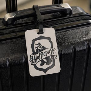 May include: A black suitcase with a Hufflepuff luggage tag. The tag is rectangular with a black and white crest featuring a badger and the word "Hufflepuff" on a banner. The luggage tag is attached to the suitcase handle.