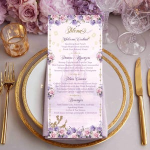 May include: A decorative menu card with a floral design in purple and pink, with a gold border. The menu lists cocktails, starters, main courses, and drinks. The card is on a white plate with gold trim, with gold cutlery and a glass.
