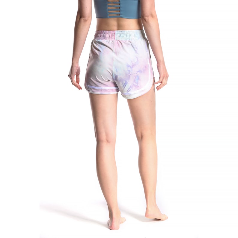 May include: A pair of white athletic shorts with a tie-dye pattern in shades of pink, blue, and purple. The shorts have a white elastic waistband and white piping along the sides.