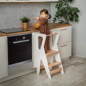 May include: A white wooden learning tower with two steps, a child is standing on the top step holding a wooden spoon and a bowl. The tower is in a kitchen setting with white cabinets and a black oven.