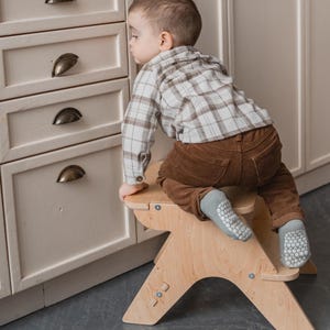 Puede incluir: Un taburete de madera de color claro con un diseño único. Un niño está usando el taburete para alcanzar un armario. El niño lleva una camisa a cuadros, pantalones marrones y calcetines grises. El armario tiene tiradores de metal oscuro.