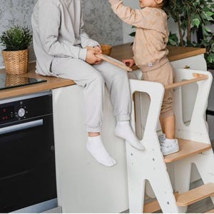 Puede incluir: Una torre de aprendizaje de madera blanca con diseño de taburete, que cuenta con paneles laterales con asas recortadas y una barra de seguridad horizontal. La estructura está construida con madera clara y paneles pintados de blanco.