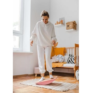 Puede incluir: Una tabla de equilibrio de madera curvada con la parte inferior de color rosa claro y la superficie superior de madera natural. La tabla está colocada sobre una alfombra tejida rectangular y presenta un diseño arqueado y liso.