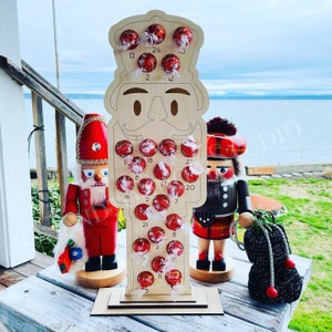 Può includere: Calendario dell'avvento in legno a forma di schiaccianoci con 24 scomparti numerati pieni di caramelle al cioccolato rosso e bianco. Il calendario è a forma di schiaccianoci con un cappello rosso e bianco e una barba nera. Due piccole figurine di schiaccianoci si trovano su entrambi i lati del calendario.