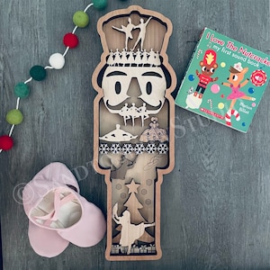 May include: A wooden nutcracker ornament with a detailed design of a Christmas tree, snowflakes, and dancers. The nutcracker is painted in brown and white colors. The ornament is a great addition to any Christmas decor.