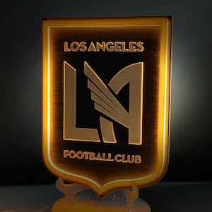 May include: A lighted Los Angeles Football Club emblem. The shield-shaped sign features the team's logo in gold against a black background, with the words "LOS ANGELES" and "FOOTBALL CLUB". The sign is mounted on a clear stand and has a warm, glowing border.