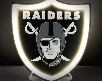 May include: A shield-shaped LED light displaying the Raiders logo. The logo features a pirate with an eye patch, crossed swords, and the word "RAIDERS" in white. The light has a white border and a black background.