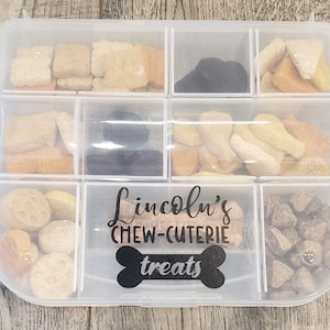 May include: A clear plastic container with multiple compartments filled with different types of dog treats. The container has a label that says "Lincoln's Chew-Cuterie treats".