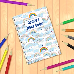Könnte beinhalten: Ein spiralgebundenes Notizbuch mit einem weißen Einband, der mit einem bunten Regenbogenmuster und dem Text "Grace's Note Book" verziert ist. Das Notizbuch ist von Buntstiften auf einer Holzoberfläche umgeben.