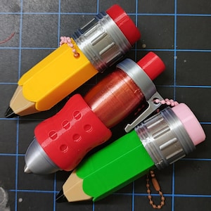 Könnte beinhalten: Drei bleistiftförmige Behälter mit Kappen. Einer ist gelb mit roter Kappe, einer ist metallisch rot mit silbernem Clip und roter Kappe, und einer ist grün mit rosa Kappe. Jeder hat eine beige Spitze, eine silberfarbene Zwinge und eine kleine Kugelkette.