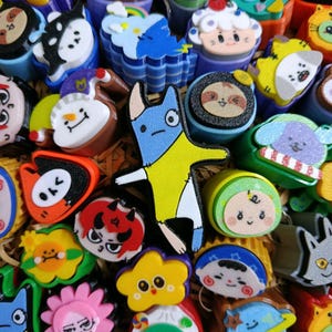 May include: A collection of colorful, small, three-dimensional plastic character stamps featuring various cartoon faces, animals, and shapes, including a central blue and yellow creature, all arranged closely together in a dense, top-down view.