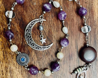 May include: A purple and white beaded pendulum with a crescent moon charm, a star charm, a blue gemstone charm, and a brown stone charm. The pendulum is hanging from a silver chain.