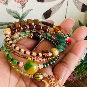 Può includere: Una collezione di braccialetti di perline nei toni del verde, marrone e oro. I braccialetti presentano varie perline, tra cui cubi di legno, perline rotonde e dettagli a forma di stella. Un braccialetto ha un ciondolo con una luna crescente e un pentagramma.