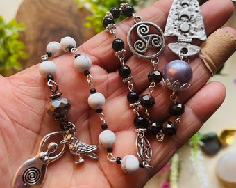 Puede incluir: Un collar de cuentas negras y blancas con dijes de plata, incluyendo una triple espiral, una luna creciente, un pentagrama, un cuervo y una mujer con una espiral dentro.