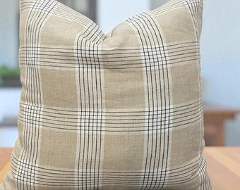 May include: A square decorative pillow with a beige background and a plaid pattern in black and white. The pillow is made of a soft fabric and is placed on a wooden surface.