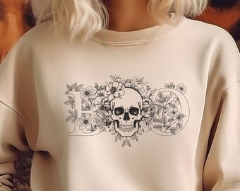 May include: A beige sweatshirt featuring a floral skull design with the word "Love" in a decorative font. The graphic is in black and white. The sweatshirt has a crew neck and long sleeves.