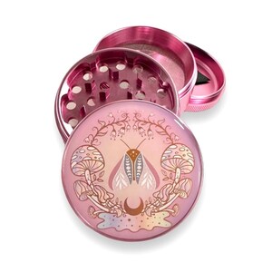 May include: A pink metal grinder with a floral design featuring a moth and mushrooms. The grinder has a three-piece design with a screen and a herb catcher.