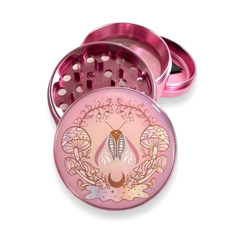 May include: A pink metal grinder with a floral design featuring a moth and mushrooms. The grinder has a three-piece design with a screen and a herb catcher.