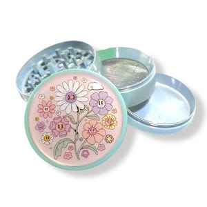 May include: A disassembled, iridescent grinder with a floral design. The top piece features a light pink background with cartoon flowers in various colors. The grinder is made of metal and has a mesh screen and a grinding chamber.