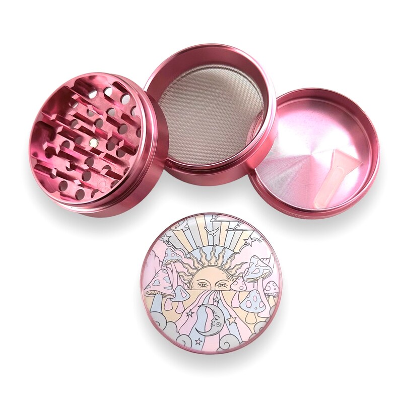 Peut inclure: Un broyeur &agrave; herbes en m&eacute;tal rose d&eacute;mont&eacute;. Le broyeur a un tamis, un petit grattoir et un couvercle avec une illustration color&eacute;e d'un soleil, de champignons et d'un croissant de lune. Le broyeur est con&ccedil;u pour les herbes.