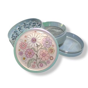 May include: A four-piece herb grinder with a pastel color scheme. The top piece features a floral design with smiling flowers in pink, purple, and yellow. The grinder is made of metal and has a mesh screen.