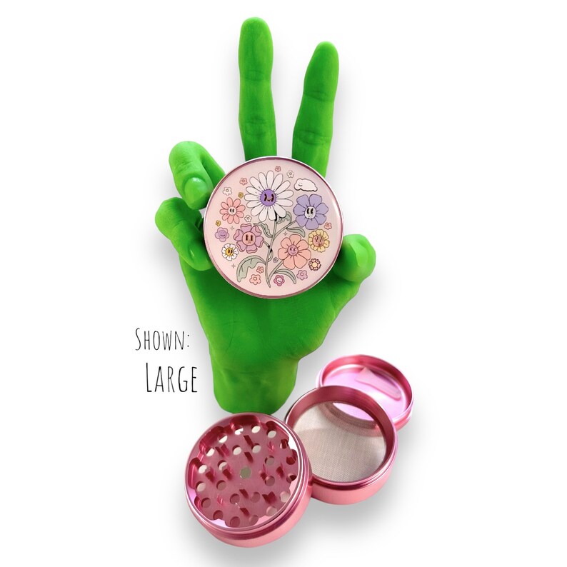 May include: A pink, three-piece grinder with a floral design. The top piece features a colorful flower illustration. The grinder is shown with the text "SHOWN: LARGE".