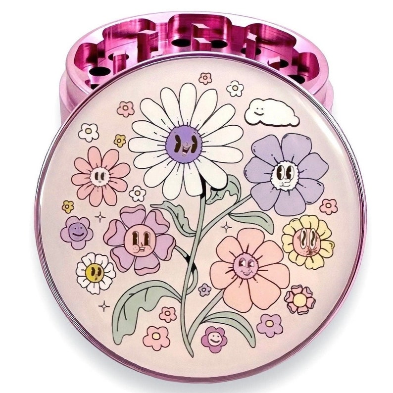 May include: A pink metal herb grinder with a floral design. The top of the grinder features a clear, circular design with cartoon flowers in pastel colours. The grinder's base is a solid pink colour.