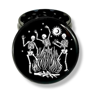 May include: Black round grinder with a white illustration of three skeletons dancing around a bonfire under a crescent moon and stars. The grinder is made of metal and has a two-piece design.