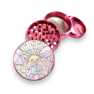 Peut inclure: Un broyeur en m&eacute;tal rose d&eacute;mont&eacute; avec un design fantaisiste. La partie sup&eacute;rieure pr&eacute;sente une illustration color&eacute;e d'un soleil, de champignons et d'un croissant de lune. Le broyeur est con&ccedil;u avec plusieurs compartiments et un tamis.