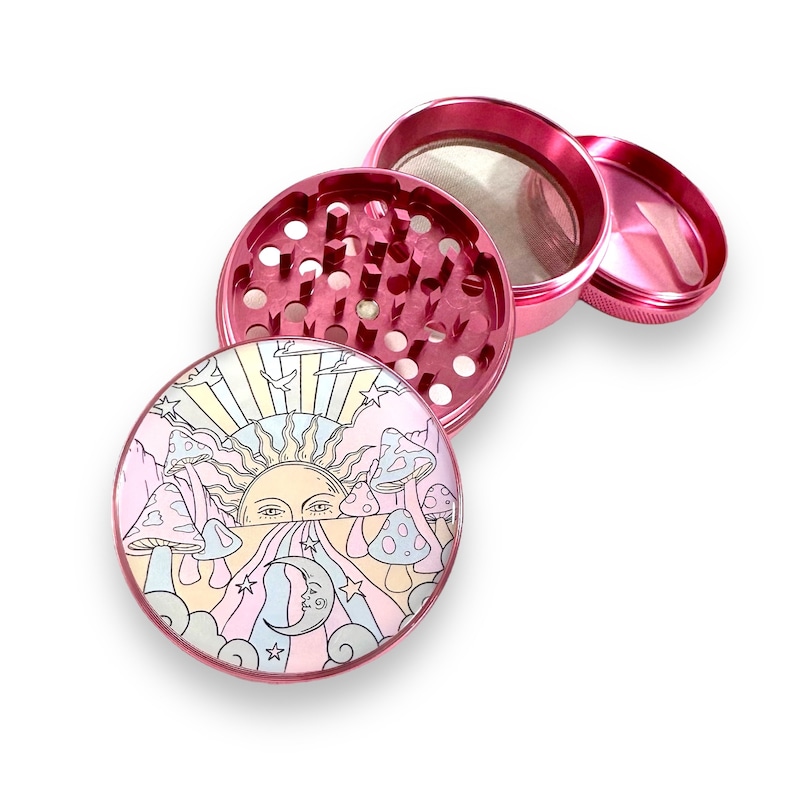 Peut inclure: Un broyeur en m&eacute;tal rose d&eacute;mont&eacute; avec un design fantaisiste. La partie sup&eacute;rieure pr&eacute;sente une illustration color&eacute;e d'un soleil, de champignons et d'un croissant de lune. Le broyeur est con&ccedil;u avec plusieurs compartiments et un tamis.