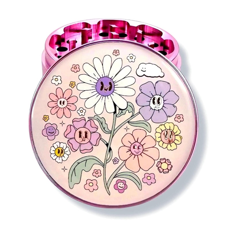 May include: A pink and silver herb grinder with a whimsical floral design. The top of the grinder features a series of teeth, while the bottom has a colourful illustration of smiling flowers in various colours, including white, pink, and purple.