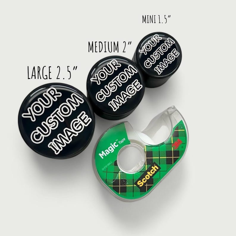 Pode incluir: Uma variedade de objetos redondos pretos com o texto "YOUR CUSTOM IMAGE" em branco, em tr&ecirc;s tamanhos: grande 6,35 cm, m&eacute;dio 5,08 cm e mini 3,81 cm. Um dispensador de fita adesiva Scotch Magic Tape xadrez verde e preto tamb&eacute;m est&aacute; presente.