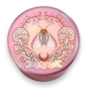 May include: A pink metal grinder with a circular design featuring a moth with moon and star accents, surrounded by mushrooms and floral elements.