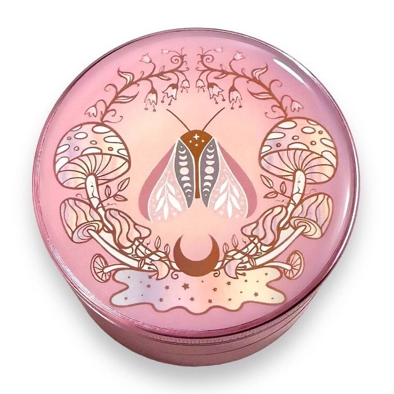 May include: A pink metal grinder with a circular design featuring a moth with moon and star accents, surrounded by mushrooms and floral elements.