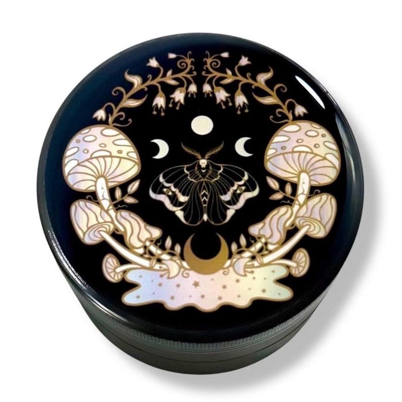 Pode incluir: Um moedor redondo preto com um design celestial. O design apresenta dois cogumelos, uma mariposa, luas crescentes e uma lua cheia. Os cogumelos e a mariposa s&atilde;o em tons de branco e dourado, sobre um fundo preto.