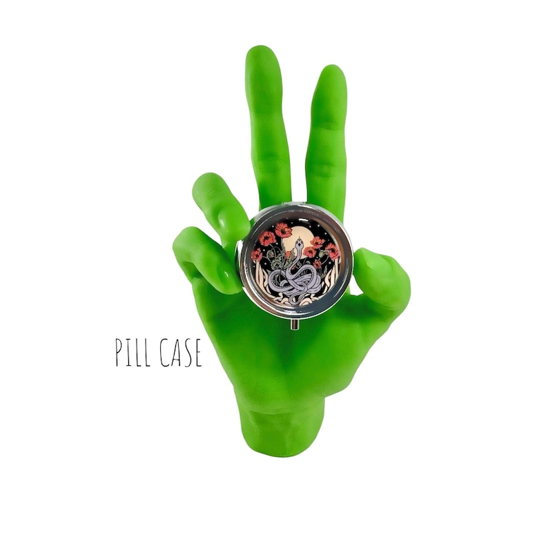 Puede incluir: Un pastillero redondo y plateado con una ilustraci&oacute;n detallada de una serpiente, flores y una luna. El estuche est&aacute; sostenido por una mano verde brillante. El texto "PILL CASE" est&aacute; debajo.