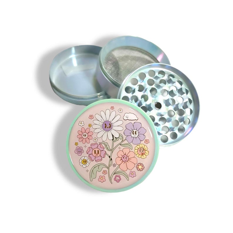 May include: A disassembled iridescent grinder with a floral design. The grinder features a light green base with a pink top adorned with cartoon flowers in various colors. The other components are silver with a mesh screen and a grinding surface.
