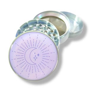Puede incluir: Un grinder de hierbas plateado con un diseño celestial. La tapa superior presenta un fondo lila claro con un diseño de luna creciente, estrella y rayos de sol. El grinder es de metal y tiene varias partes.