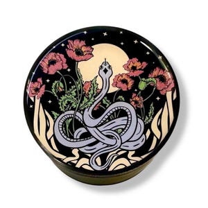 Puede incluir: Contenedor redondo y negro con una ilustraci&oacute;n detallada. Una serpiente azul claro est&aacute; enrollada frente a una luna llena, rodeada de amapolas rosas y follaje verde. El fondo es negro con estrellas.