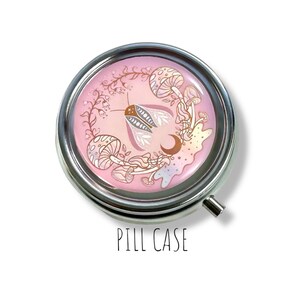 May include: A silver pill case with a pink and gold design featuring a moth, mushrooms, and a crescent moon. The text "PILL CASE" is printed below the image.